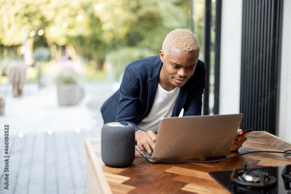 Latin man works on a laptop computer at thhe kitchen table with smart speaker. Controlling smart hom