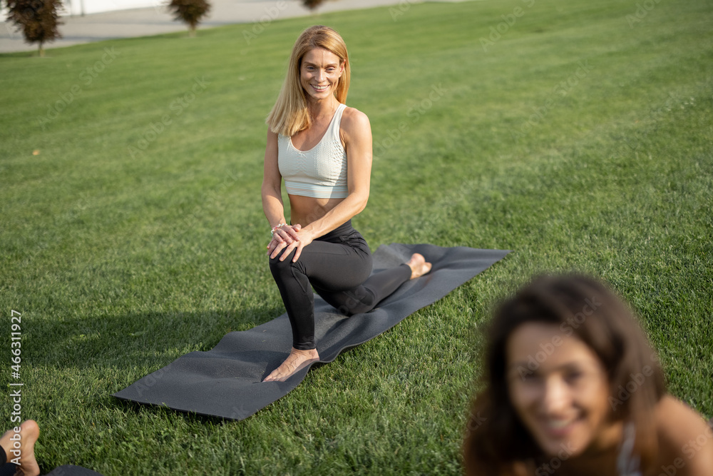 Athletic girlfriends stretching before sports training on fitness mats on green lawn at sunny day. C