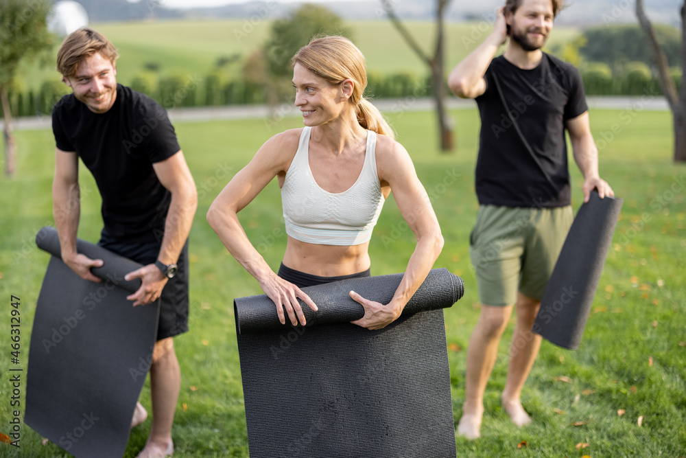 Group of pleased european people folding fitness mats after training on green meadow. Concept of hea