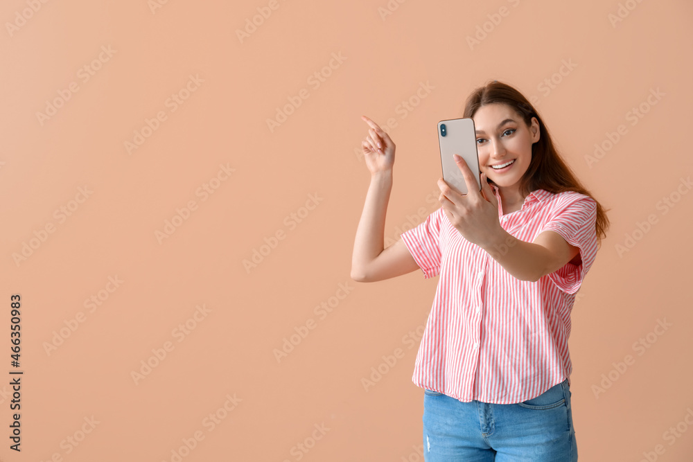 Young smiling woman taking selfie and pointing at something on beige background