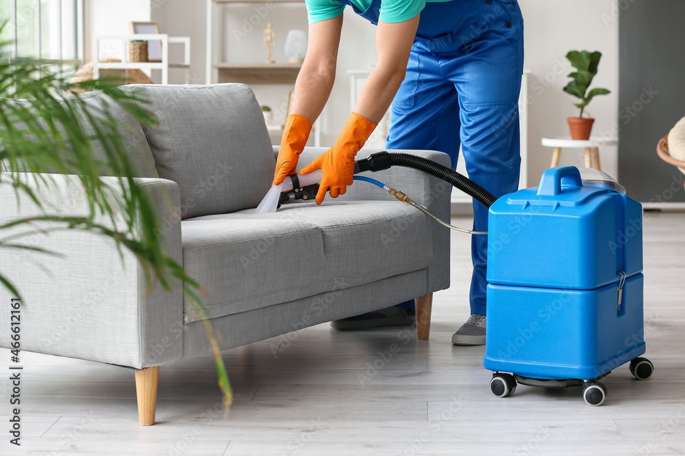 Male worker removing dirty stain from grey sofa with vacuum cleaner in room