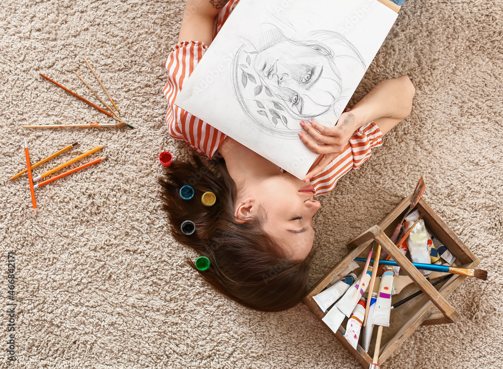 Young female artist lying on carpet at home, top view