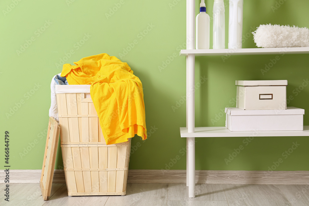 Wooden basket with dirty clothes and shelving unit near green wall