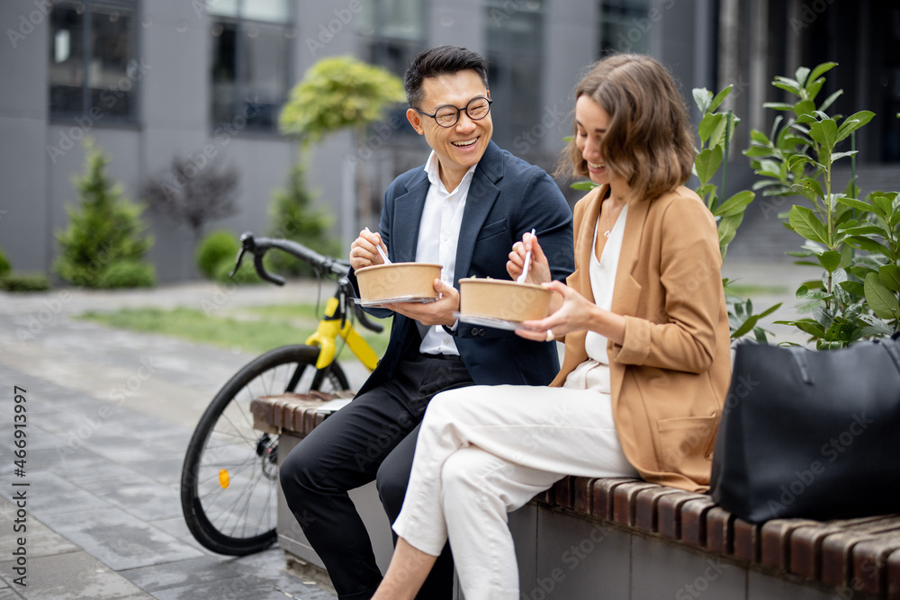 Asian businessman and caucasian businesswoman eating food and talking while having lunch at work. Co