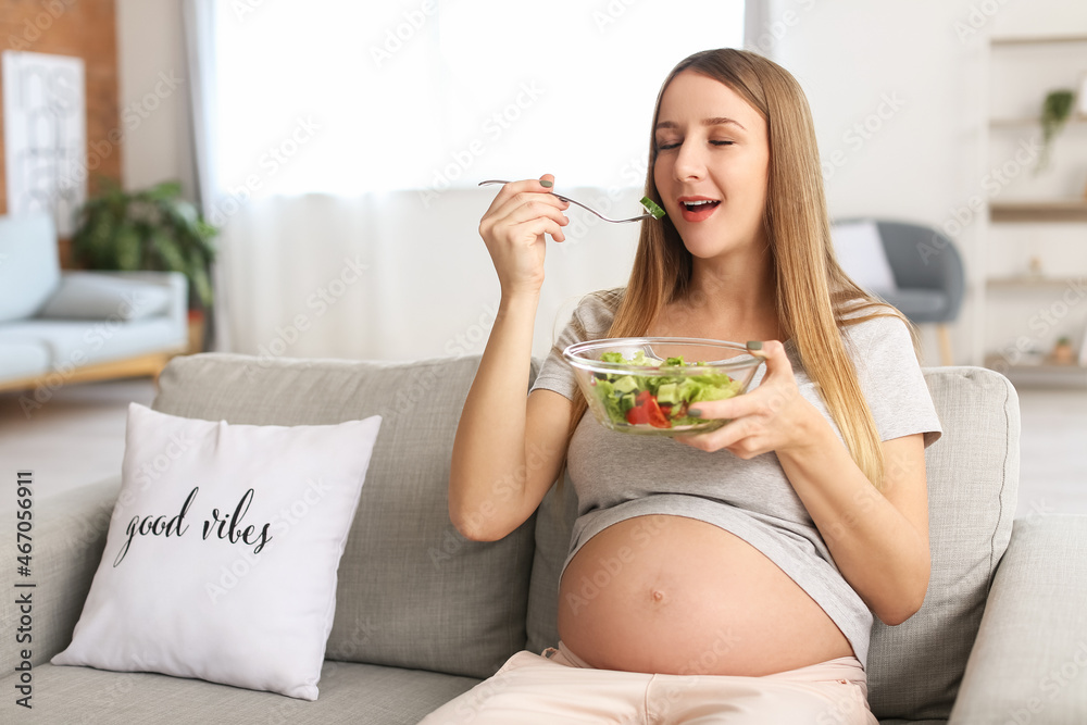 Young pregnant woman eating vegetable salad on sofa at home