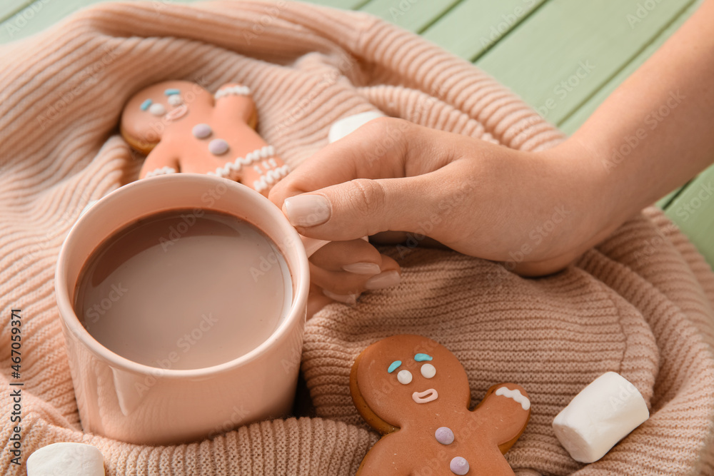 Female hand with cup of cacao, marshmallow, cookies and sweater on color wooden table, closeup