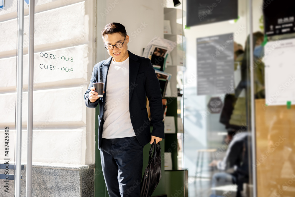 Asian businessman with coffee going from entrance of building. Smiling modern adult successful man w