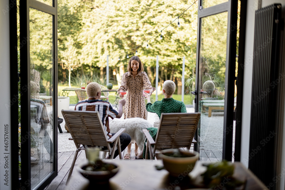 Woman giving cocktails to a friends sitting on wooden chairs at home terrace. European girls and lat