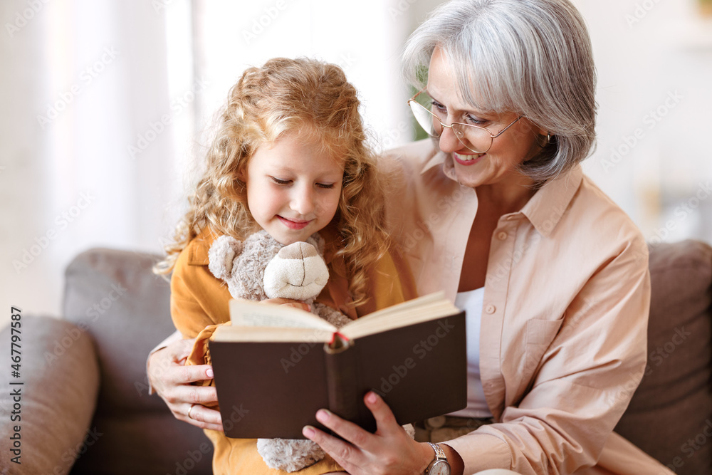 Cute little girl granddaughter reading book with positive senior grandmother