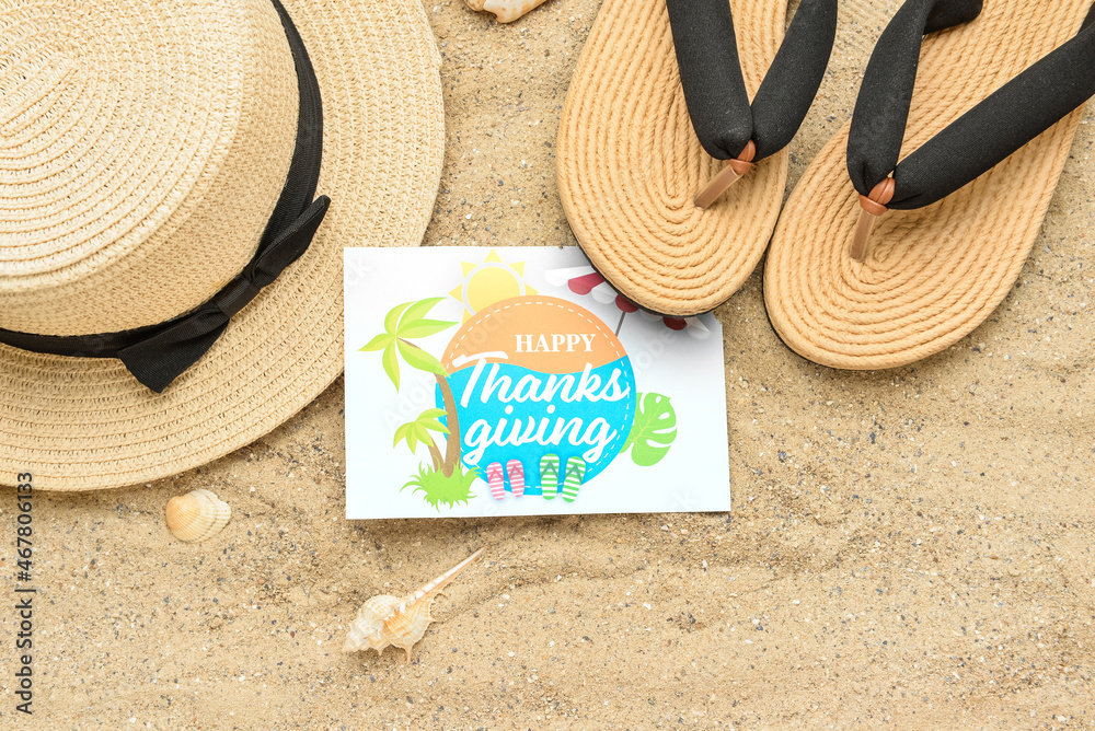 Beach hat, flip flops and card with text HAPPY THANKSGIVING on sand background