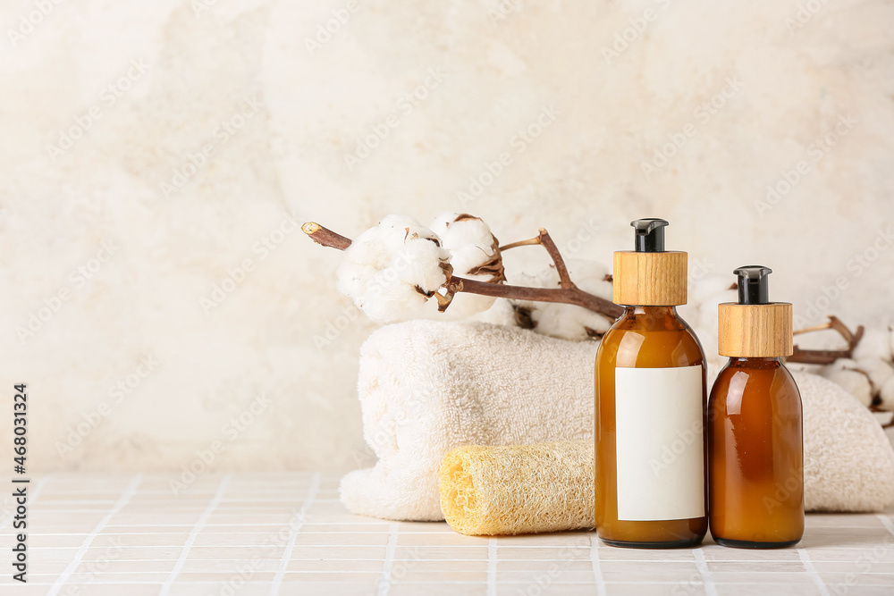 Bottles of cosmetic products, towel, sponge and cotton branch on tile table against light background