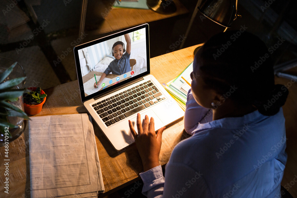 African american woman using laptop for video call, with elementary school pupil on screen