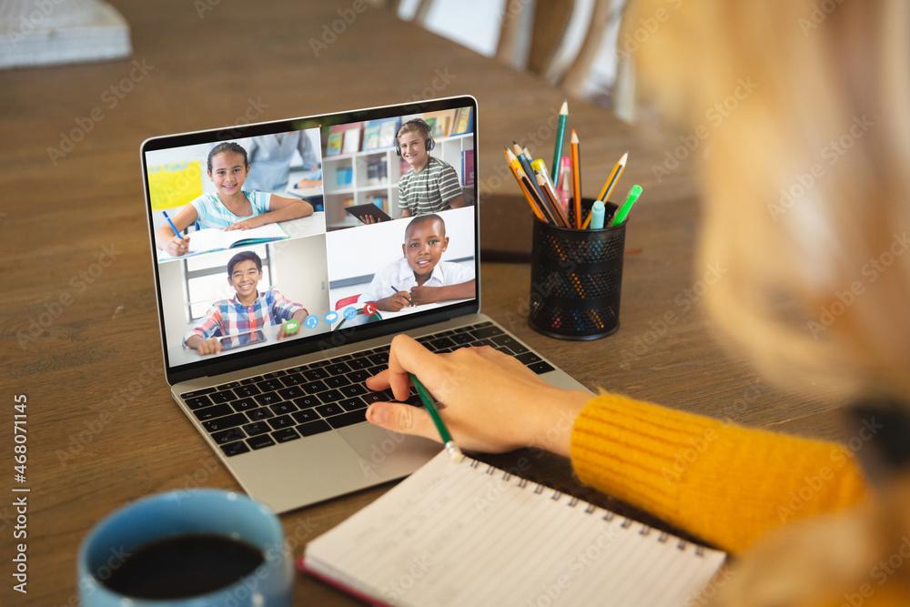 Caucasian woman using laptop for video call, with smiling diverse elementary school pupils on screen