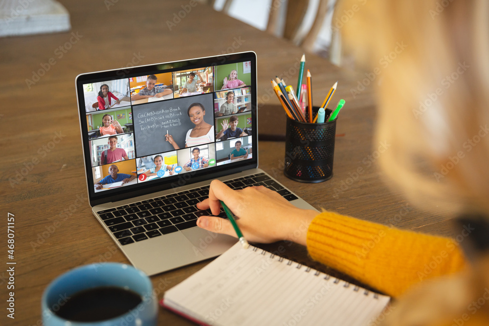 Caucasian woman using laptop for video call, with smiling diverse elementary school pupils on screen