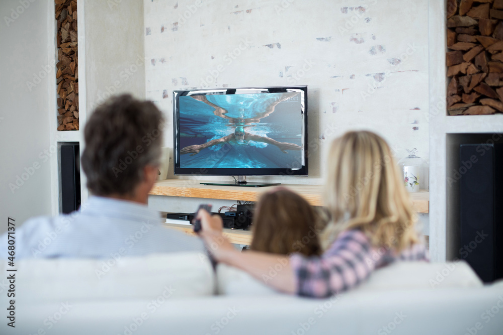 Rear view of family sitting at home together watching swimming competition on tv