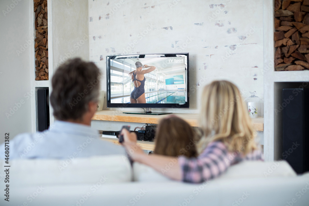 Rear view of family sitting at home together watching swimming competition on tv