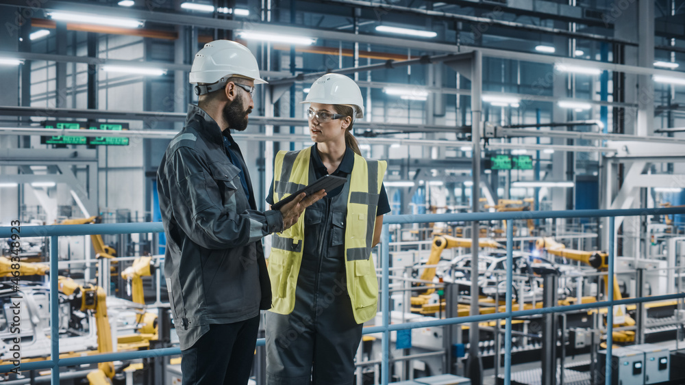 Male Specialist and Female Car Factory Engineer in High Visibility Vests Using Tablet Computer. Auto