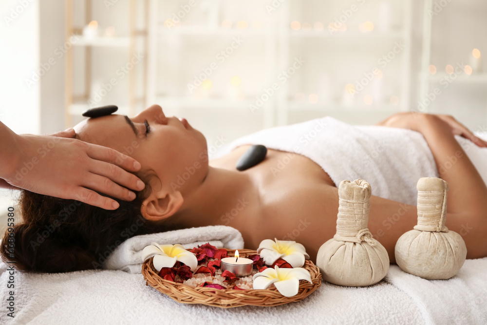 Young African-American woman getting facial massage by therapist with stones in spa salon