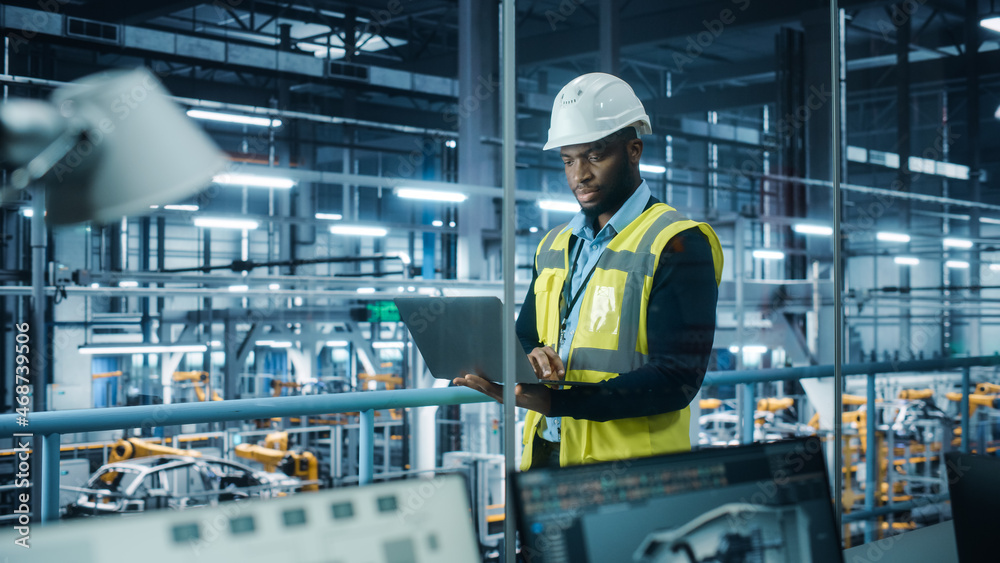 Car Factory: Professional Male Automotive Engineer Wearing Hard Hat, Walking, Using Laptop. Monitori