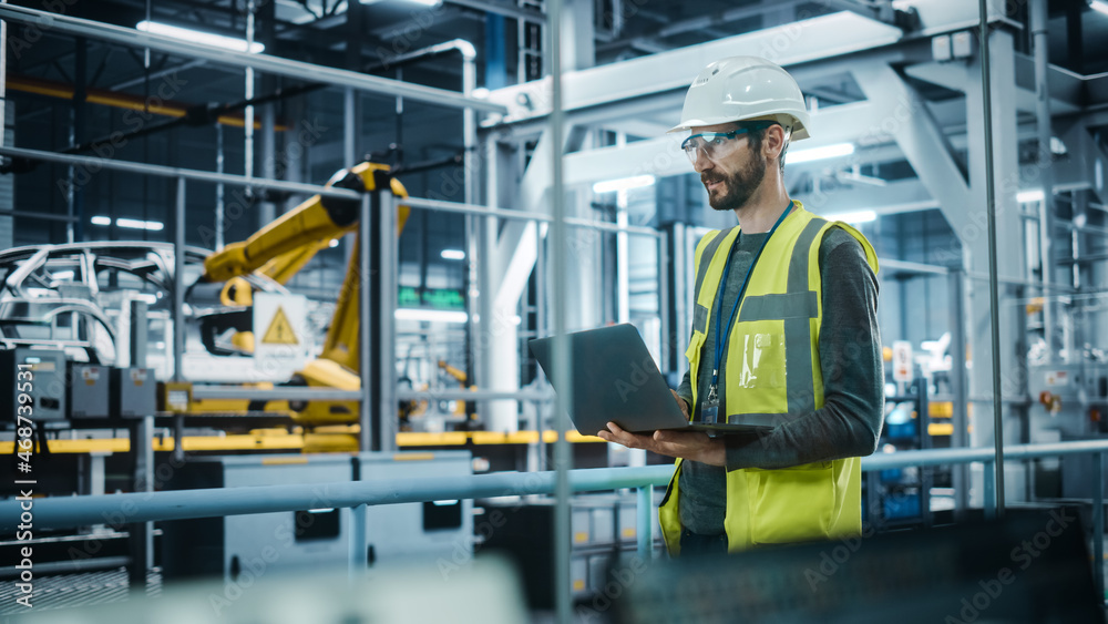 Car Factory: Male Automotive Engineer Wearing Hard Hat, Standing, Using Laptop. Monitoring, Control,