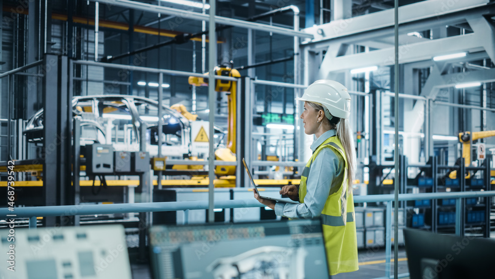 Car Factory: Female Automotive Engineer Wearing Hard Hat, Standing, Using Laptop. Monitoring, Contro