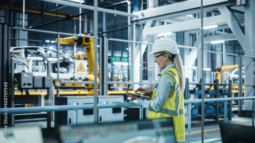 Car Factory: Female Automotive Engineer Wearing Hard Hat, Standing, Using Laptop. Monitoring, Contro