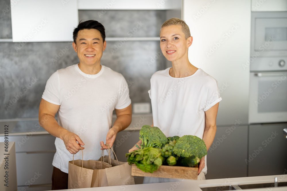 亚洲男人拿着购物袋，女人在家里的厨房里拿着新鲜蔬菜。多种族夫妇