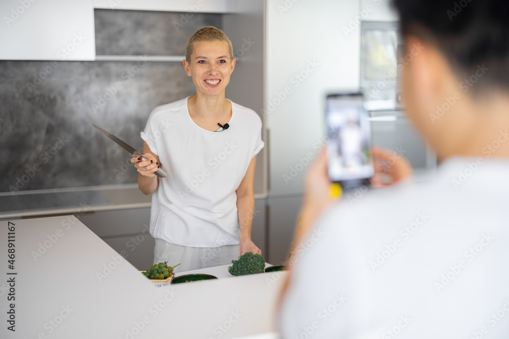 一名男子在智能手机上拍摄高加索女子在厨房烹饪素食沙拉的视频。vi的概念