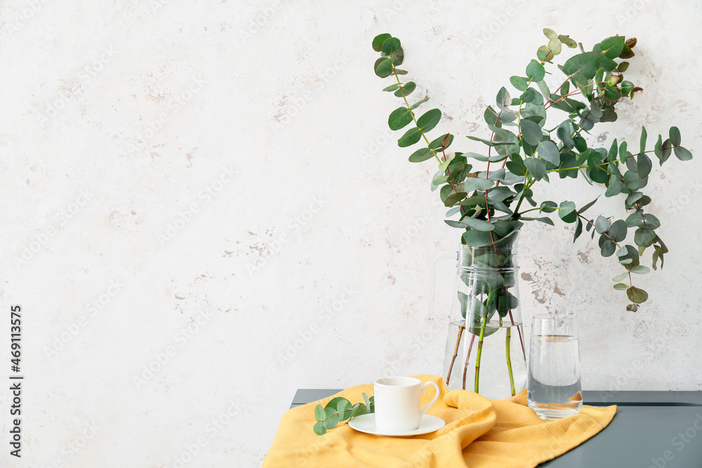 Eucalyptus branches in vase and cup of coffee on table near light wall