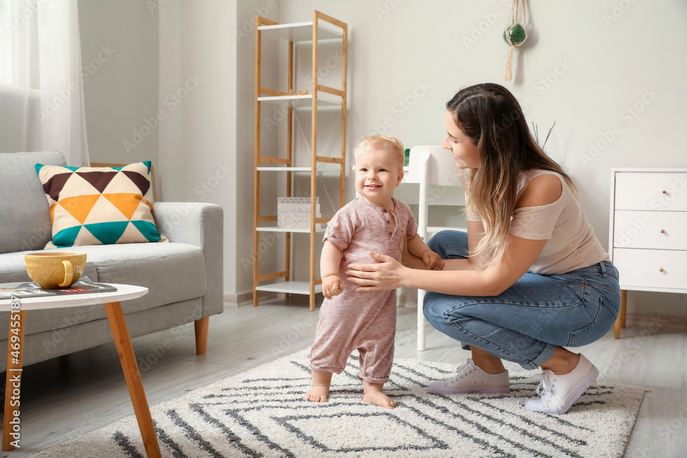 Cute baby girl in bodysuit learning to walk with her mother at home