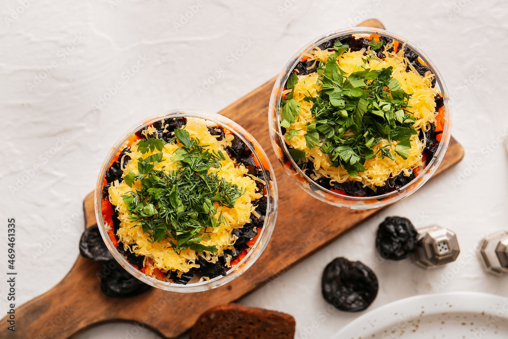 Wooden board with delicious layered salad with prunes in glasses on light background