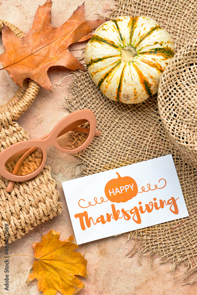 Wicker bag, hat, dry leaves, pumpkin, sunglasses and paper card with text HAPPY THANKSGIVING on colo