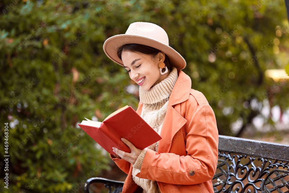 Beautiful young woman sitting on bench and reading interesting book in park