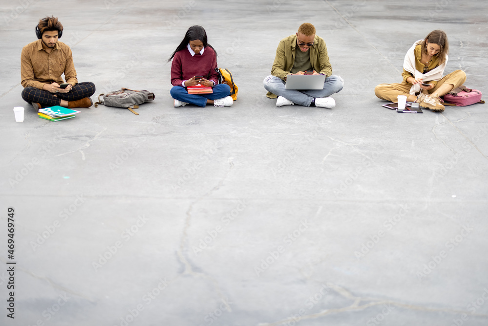 Multiracial students sitting and using gadgets on asphalt at university campus. Concept of education