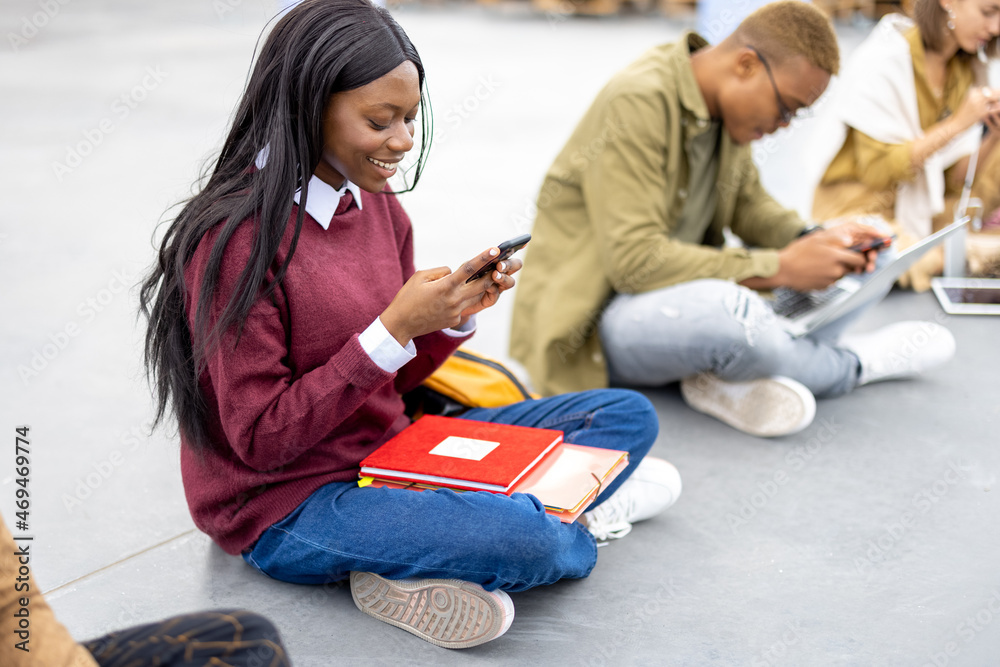 Row of multiracial students sitting and using smartphones on asphalt at university campus. Concept o