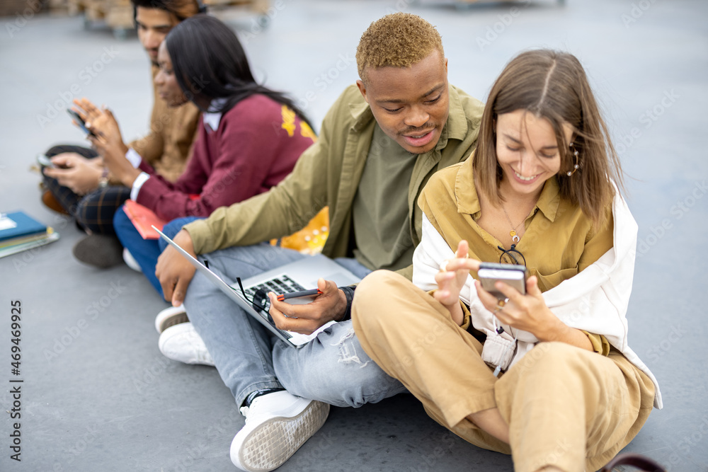 Multiracial students sitting and using modern gadgets on asphalt at university campus. Concept of ed