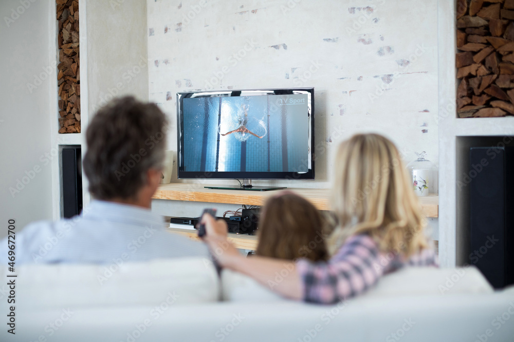 Rear view of family sitting at home together watching swimming competition on tv