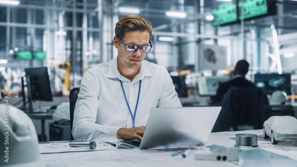 Young Handsome Engineer Working and Managing Projects on Laptop Computer in an Office at Car Assembl