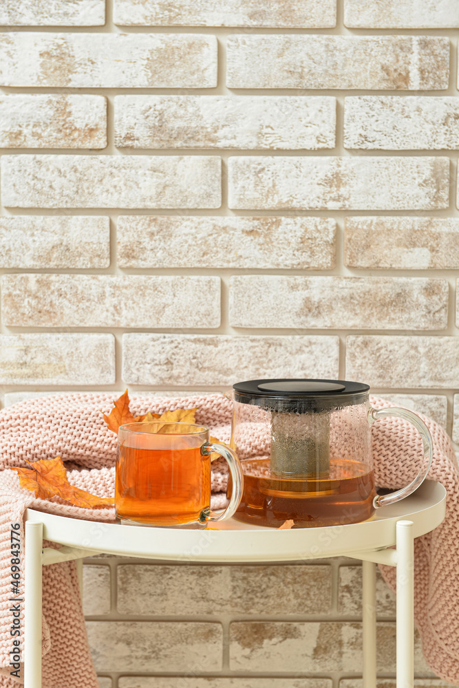 Tea pot, glass cup of hot beverage and autumn leaves on table near brick wall