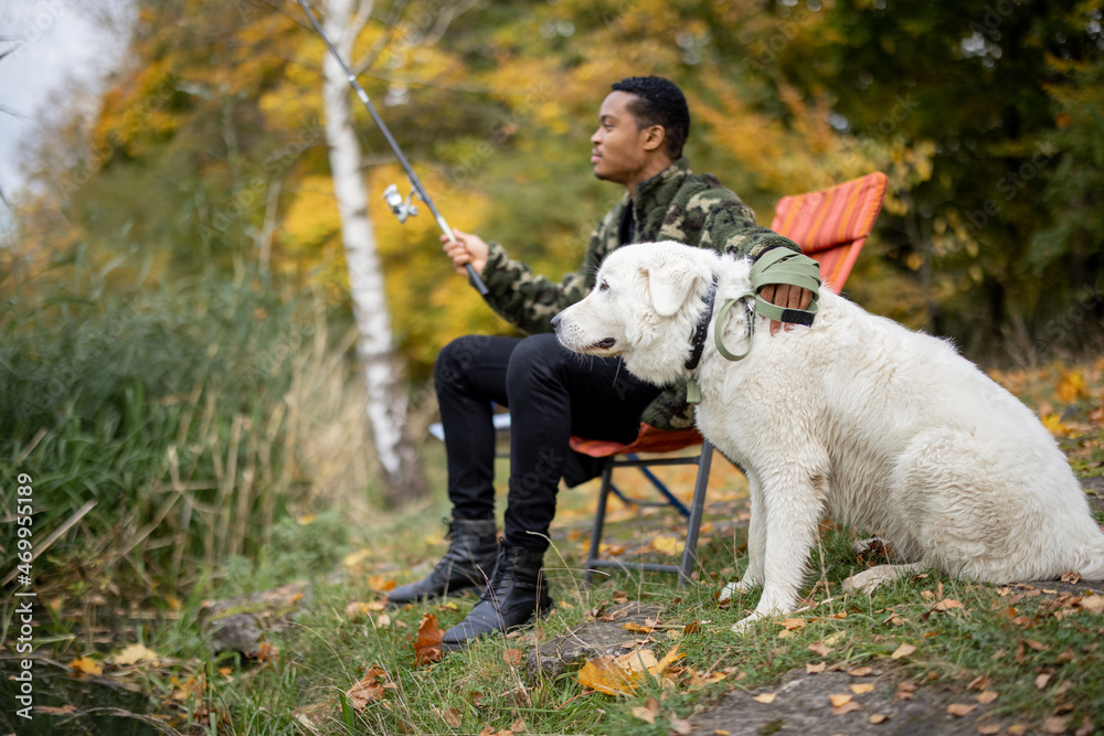 一名拉丁男子坐在椅子上，在河流或湖泊海岸的Maremmano Abruzzese Sheepdog附近钓鱼。Con