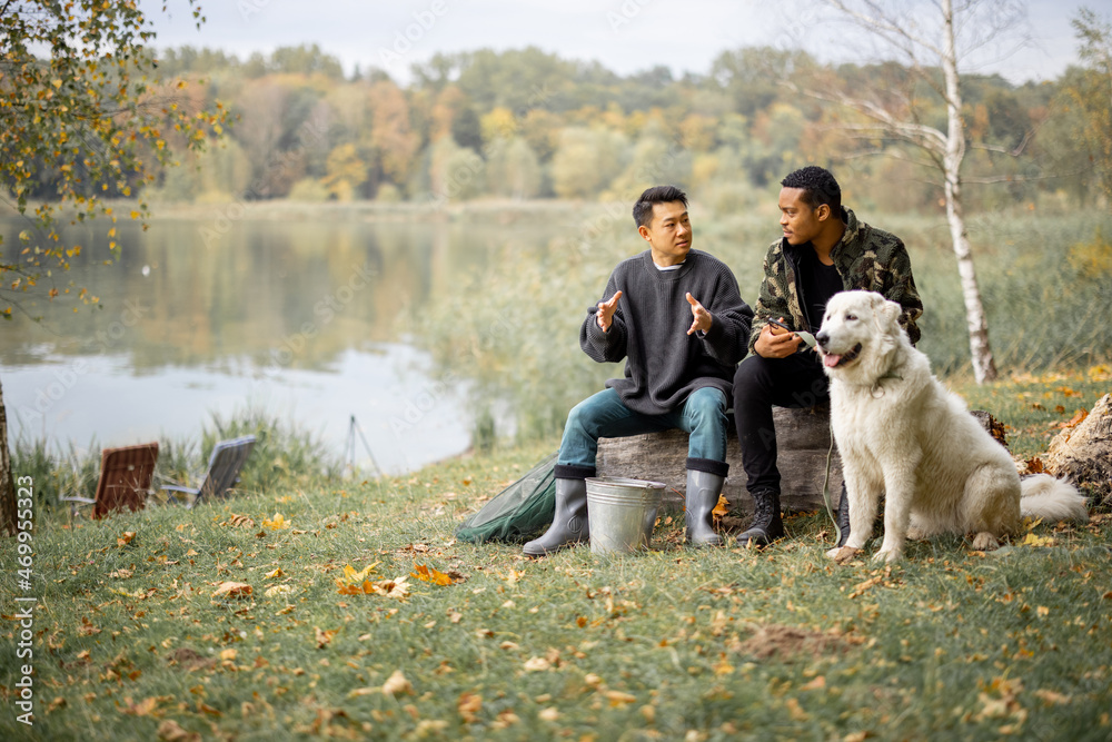 多种族男性朋友坐在Maremmano Abruzzese Sheepdog附近倒下的树上聊天