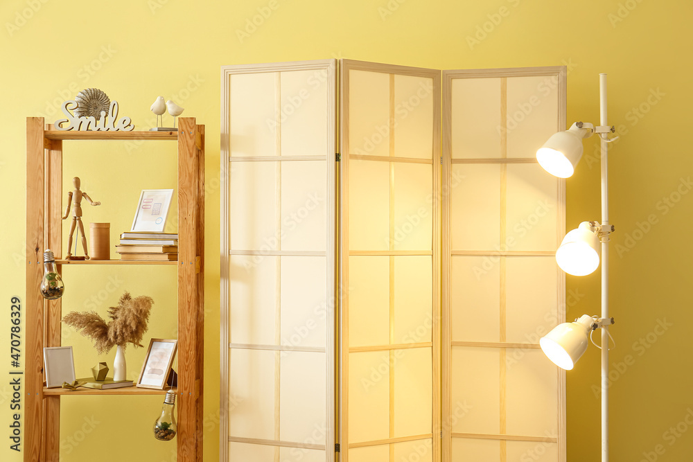 Folding screen with glowing lamp and shelving unit near yellow wall