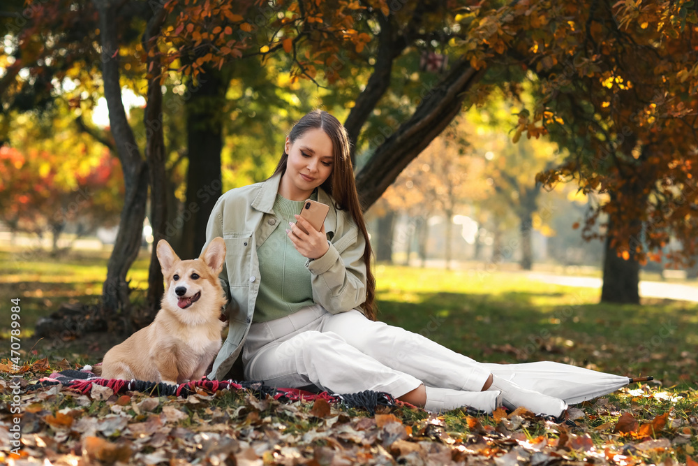 年轻女子带着可爱的柯基犬在秋季公园使用手机