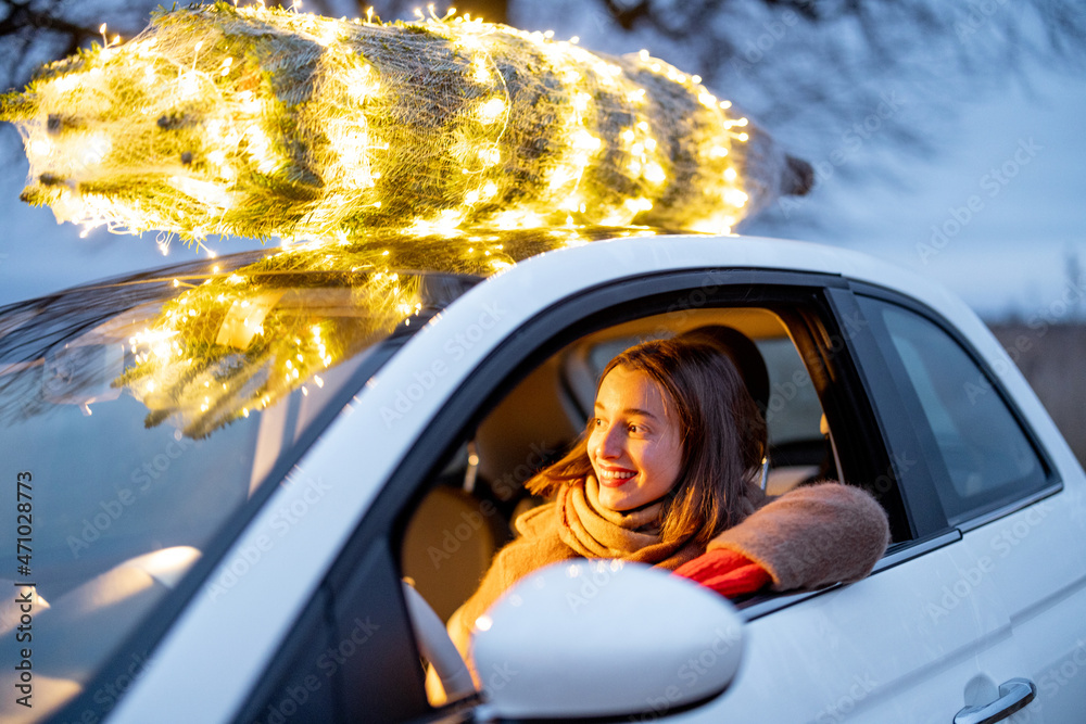 Happy woman driving car with a illuminated Christmas tree on a rooftop. Concept of New Year preparat