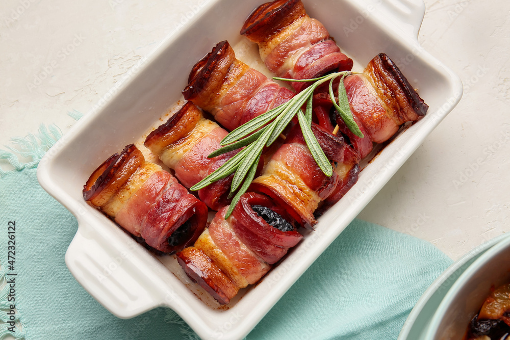 Baking dish with delicious prunes baked in crispy bacon and rosemary branch on light background, clo