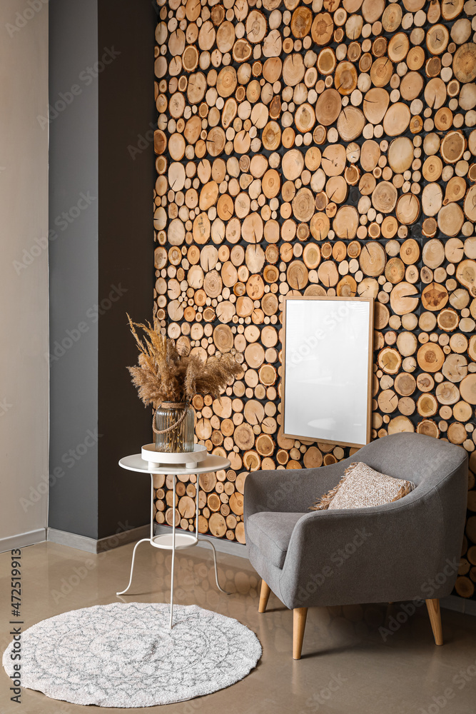 Glass vase with dry reeds on table and grey armchair near wooden wall