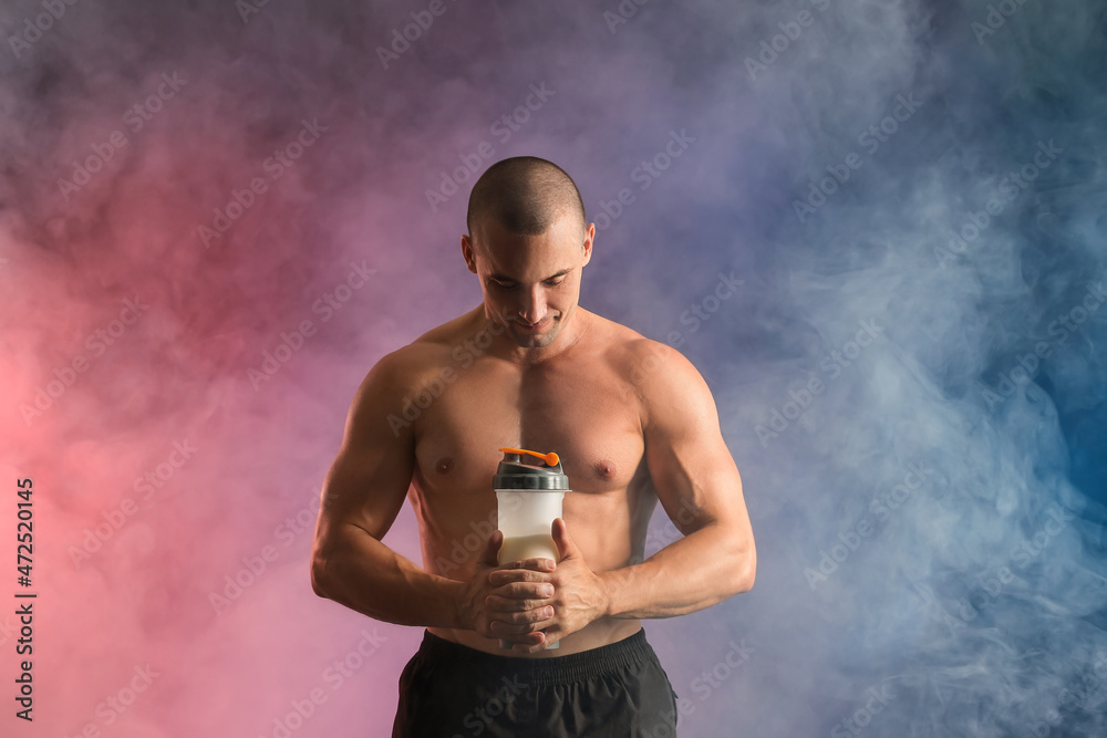 Male bodybuilder with protein shake in smoke on color background