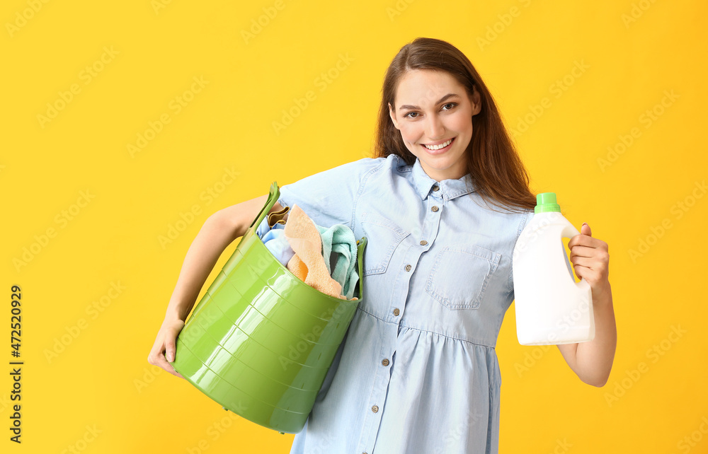 Beautiful woman with detergent and laundry in green basket on yellow background