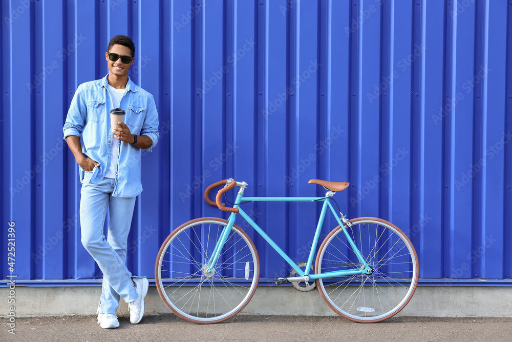 African-American teenage boy with cup of coffee and bicycle near blue fence