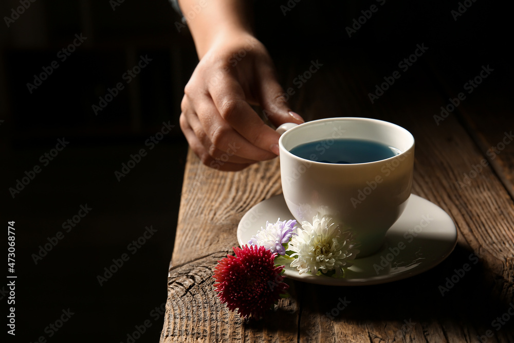 深色背景下，木桌上放着一杯蝴蝶豌豆花茶的女性手